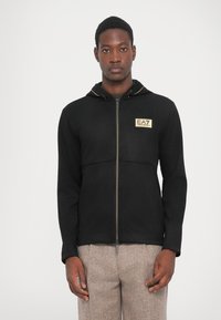 Black zip-up jacket with a hood, featuring a gold logo patch on the left side, textured fabric, and contrast stitching.
