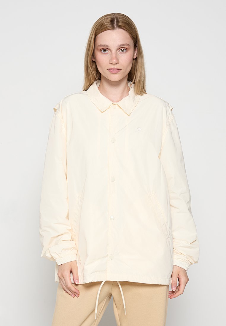 Light cream windbreaker jacket with a collared neck, snap buttons, side pockets, and elastic cuffs. Features a hem drawstring for fit adjustment.