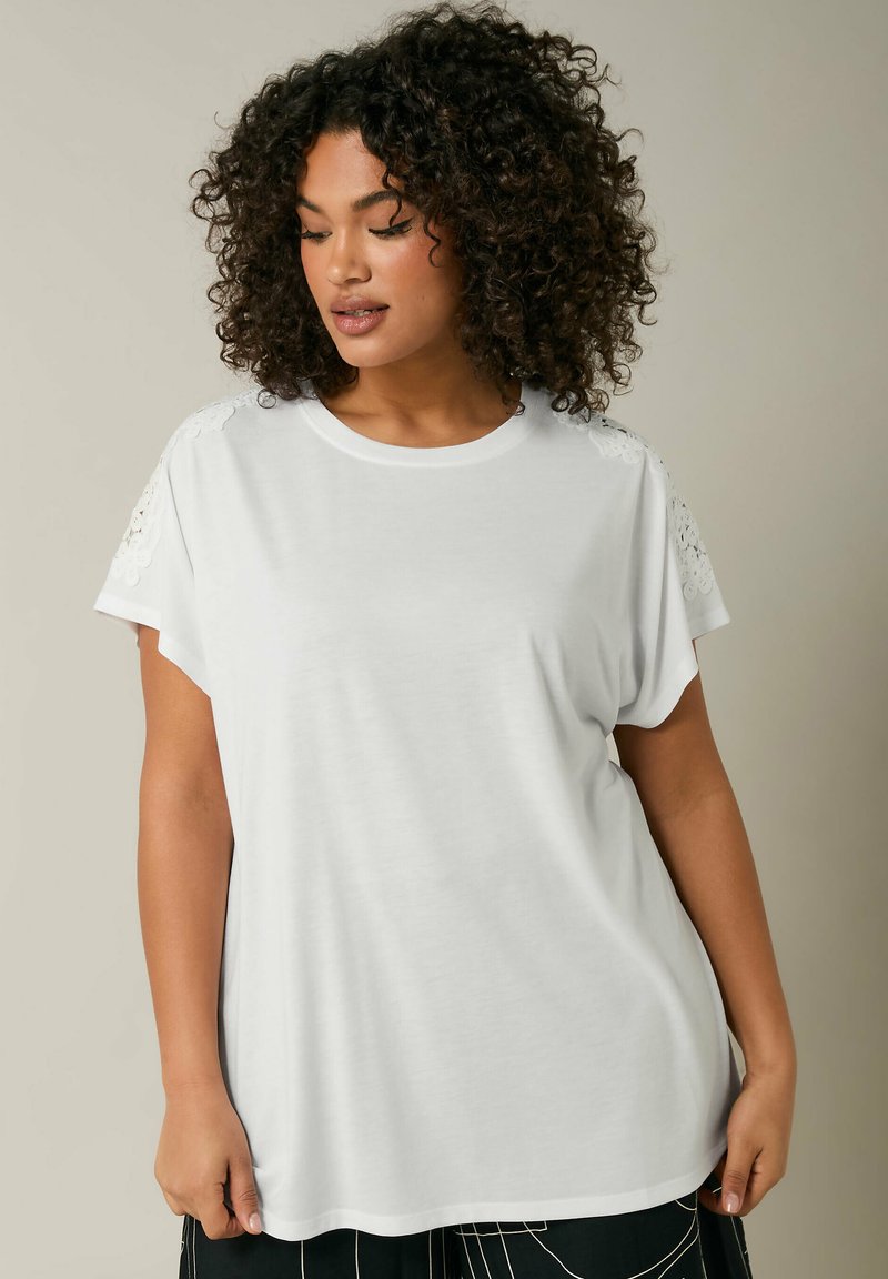 T-shirt bianco a maniche corte realizzato in un tessuto morbido, con dettagli in pizzo sulle spalle e una vestibilità rilassata e ampia.