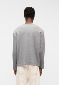 Άνδρας με κοντά σγουρά μαλλιά, φορώντας φαρδιά γκρι μπλούζα με μακριά μανίκια και ανοιχτό μπεζ παντελόνι, στέκεται με την πλάτη προς το μέρος μας σε λευκό φόντο.