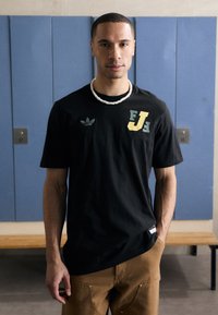 Black short-sleeve T-shirt with green and yellow graphic accents and an Adidas logo. Worn with khaki pants in a gym setting.