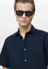 Cămașă cu mânecă scurtă, de culoare albastră închisă, cu guler, textură netedă, având butoane albe și un design minimal. Ochelari de soare negri completează ținuta.