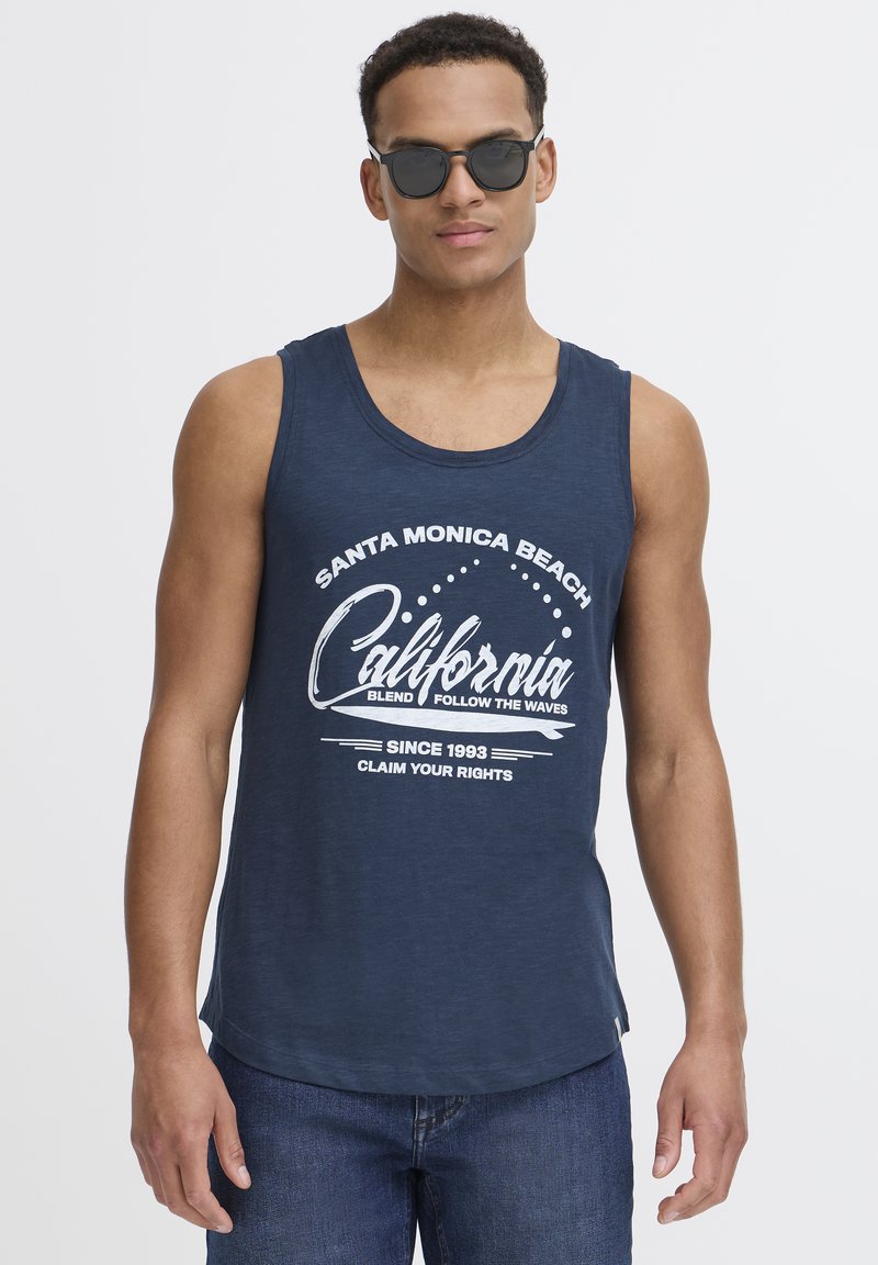 Mornarsko modra majica brez rokavov z belim grafičnim tiskom, ki vključuje besedilo "Santa Monica Beach" in "California". V kombinaciji s temnimi sončnimi očali.