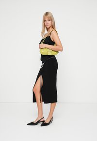 Rochie neagră cu o fanta laterală, partea superioară ajustată și detalii cu volane. Modelul ține o geantă texturată de culoare verde-lămâie, cu curașă și accente din lanț.