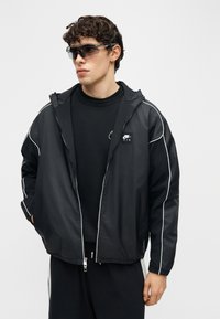 Νεαρός άνδρας που φοράει μαύρο αδιάβροχο Nike Air με άσπρη επένδυση, μαύρο πουκάμισο, σκουρόχρωμα γυαλιά ηλίου, κοιτάζει αριστερά, τα χέρια στις τσέπες.