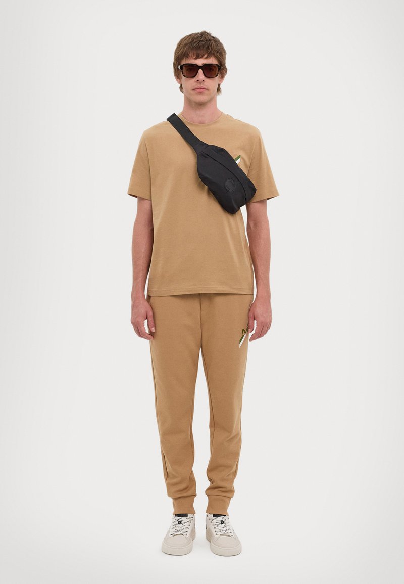 Camiseta de algodón beige y joggers a juego con un acento verde, combinados con un bolso cruzado negro y zapatillas blancas. Las gafas de sol cuadradas completan el look.