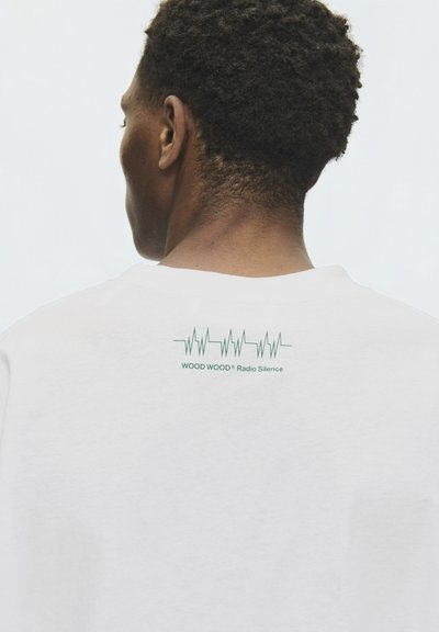 Homme aux cheveux courts et bouclés portant une chemise blanche avec un battement de cœur vert et le texte "WOOD WOOD Radio Silence" sur le haut du dos.