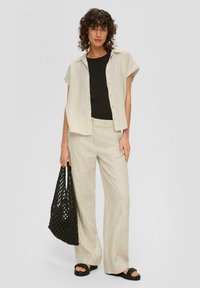 Beige linnen overhemd met korte mouwen over een zwarte tanktop, gecombineerd met lichtbeige linnen broek. Model houdt een zwarte netzak vast. Zwarte sandalen zichtbaar.
