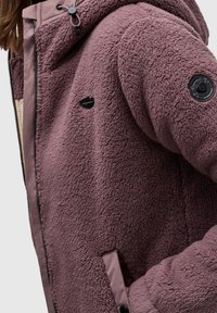 Chaqueta de forro polar difusa en color malva con capucha, que cuenta con accesorios negros, un forro interior contrastante y bolsillos laterales para mayor funcionalidad.