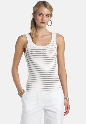 Młoda kobieta ubrana w biały, bezrękawnik prążkowany z cienkimi, poziomymi beżowymi paskami oraz białe spodnie, ozdobiona kolczykami kołami i bransoletkami.