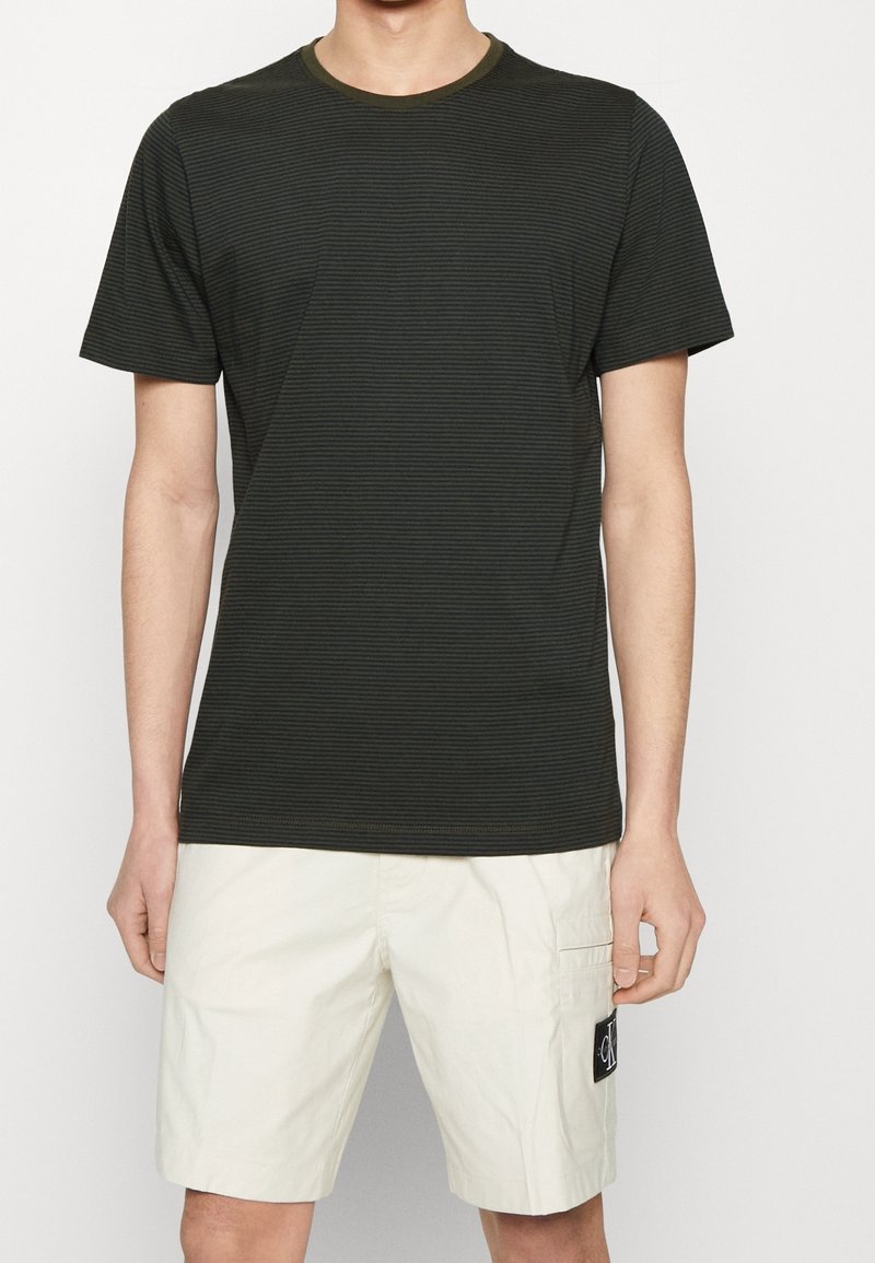 T-shirt a righe nere con colletto verde, abbinato a pantaloni corti color crema con tasca cargo e patch logo. Materiale misto cotone.