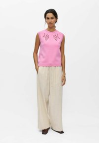 Mujer de pie, con una camiseta sin mangas rosa con patrones de hojas recortadas, pantalones beige de pierna ancha y zapatos marrón oscuro sobre un fondo blanco.
