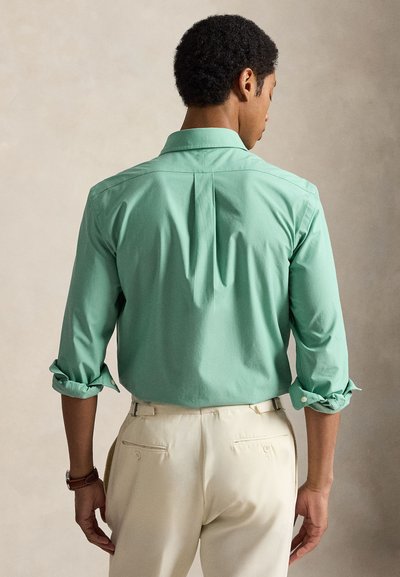 Mand vendt væk iført en opkørt mintgrøn langærmet skjorte og beige bukser med baglommer og justeringsstropper i siden.