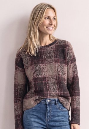 Femme blonde souriante regardant vers la droite, portant un pull à carreaux violet et marron et un jean bleu sur un fond clair.