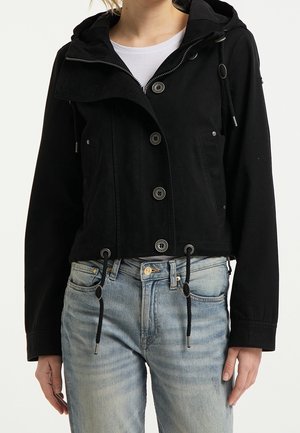 Schwarze kurzgeschnittene Jacke mit Kapuze, vorderen Knöpfen, zwei Seitentaschen und Kordelzugsaum. Getragen über einem weißen Hemd und kombiniert mit hellblauen Jeans.