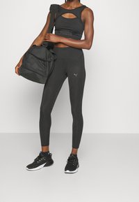 Conjunto atlético negro que incluye una camiseta sin mangas con corte y leggings de cintura alta, complementado con una bolsa de gimnasio negra y zapatillas negras.