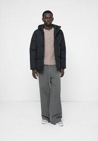 Mann iført svart pufferjakke, beige henley-skjorte, brede grå jeans og hvite joggesko som står mot en hvit bakgrunn.