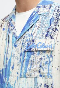Camisa de manga corta en tela ligera con un patrón de salpicaduras en azul y blanco. Cuenta con un bolsillo parche con la etiqueta "Carlo Colucci".