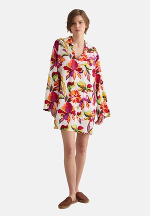Mujer con un vestido blanco suelto con grandes estampados florales de colores y zapatos marrones sin cordones, de pie con expresión neutral.