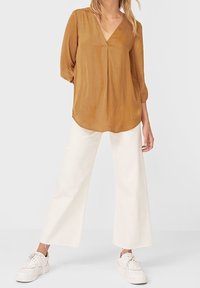 Blusa giallo senape con scollatura a V e maniche a tre quarti, abbinata a pantaloni bianchi a gamba larga e sneakers bianche.