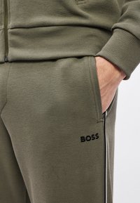 Πράσινο ελιάς τζόγκερ με υφή, σε στενή γραμμή. Διαθέτουν πλαϊνές τσέπες και μαύρο κεντημένο λογότυπο "BOSS" με λευκές πλαϊνές ρίγες.