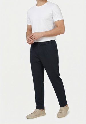 Uomo che indossa una maglietta bianca a maniche corte, pantaloni neri e scarpe da ginnastica beige, in piedi contro uno sfondo chiaro e uniforme.