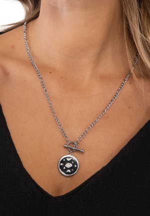 Silberne Halskette mit einem runden schwarzen Anhänger, der Mondphasen und Sterne zeigt, getragen von einer Person mit heller Haut und schwarzem Oberteil.