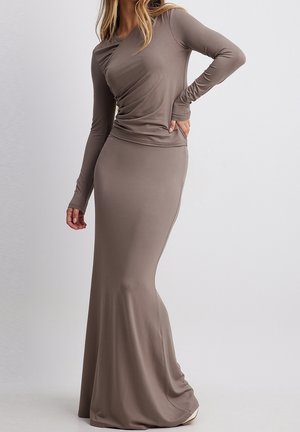 Femme portant une longue robe taupe ajustée à manches longues, debout devant un fond clair uni avec une main sur la hanche.