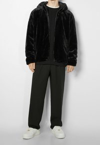 Felpa nera in pelliccia sintetica con zip, vestibilità comoda, texture morbida. Abbinata a pantaloni scuri e sartoriali e sneaker bianche. Nessun logo visibile.