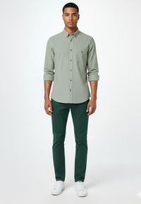 Joven de pie, vistiendo una camisa verde claro abotonada con las mangas remangadas, pantalones verde oscuro y zapatillas blancas sobre un fondo blanco liso.