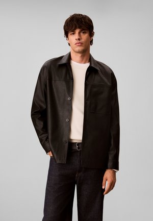 Jeune homme aux cheveux ondulés foncés portant une veste-chemise en cuir noire sur un pull blanc et un jean foncé, sur un fond uni gris clair.