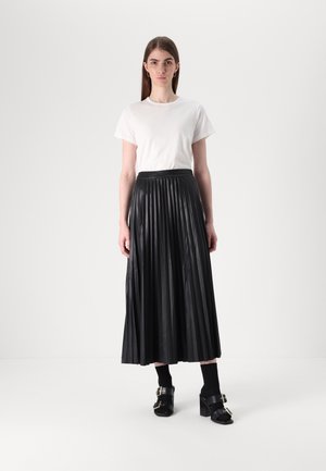 Black pleated maxi skirt with a smooth texture, paired with a white t-shirt. Model wears black chunky heels and socks, standing against a neutral background.