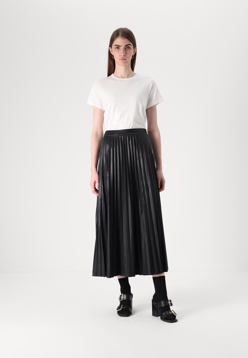 Jupe maxi plissée noire avec une texture lisse, associée à un t-shirt blanc. Le modèle porte des talons épais noirs et des chaussettes, se tenant devant un fond neutre.