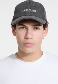 Gray denim cap with a curved brim and white "GUESS JEANS" embroidered logo. Worn with a white crewneck shirt, featuring smooth texture.