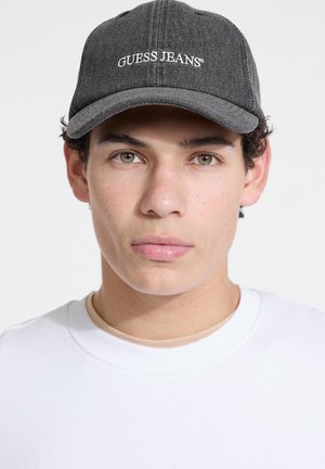Gray denim cap with a curved brim and white "GUESS JEANS" embroidered logo. Worn with a white crewneck shirt, featuring smooth texture.