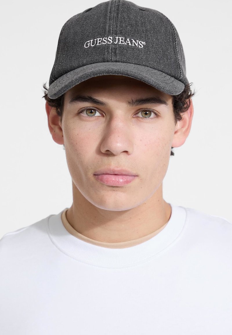 Gray denim cap with a curved brim and white "GUESS JEANS" embroidered logo. Worn with a white crewneck shirt, featuring smooth texture.