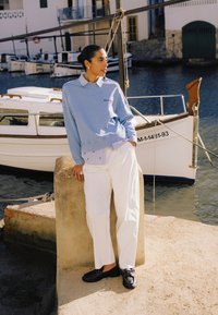 Ženska v svetlo modri majici in belih hlačah se naslanja na kamniti steber blizu privezanega čolna v mirnem pristanišču.