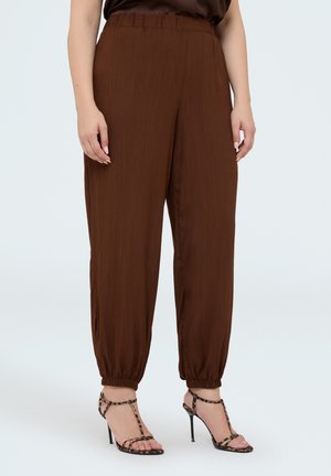 Femme portant un pantalon marron ample avec des chevilles élastiques et des sandales à talons hauts à imprimé léopard, debout devant un fond uni.