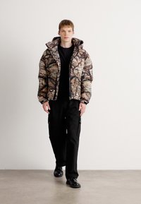 Pufferjakke med et multicolor abstrakt camouflagemønster, lynlås lukning og en tætsiddende hætte. Bæres med sorte cargo-bukser og sko.