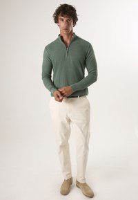 Grüner Zip-Pullover, cremefarbene Hose und beigefarbene Schuhe. Der Pullover hat einen hohen Kragen und eine figurnahe Passform mit strukturiertem Stoff.