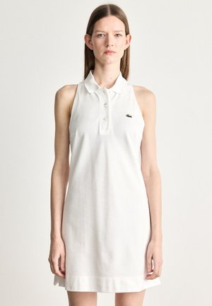 Vrouw in een mouwloze witte pol dresses met knopen en een klein groen geborduurd logo op de borst, staand tegen een effen lichte achtergrond.