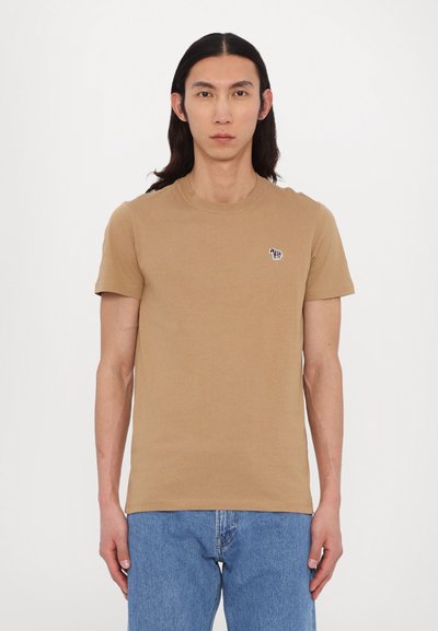 T-shirt en coton marron avec un col rond et des manches courtes ; présente un petit design brodé sur la poitrine. Accompagné d'un jean bleu.