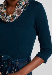 Långärmad topp i mörk teal med en dekorativ, mönstrad cowl-krage och ett tygbälte med färgglada cirkulära detaljer.