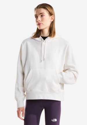 Młoda kobieta w białej bluzie z kapturem i kieszenią z przodu oraz fioletowych legginsach z logo The North Face, patrząca lekko w bok.