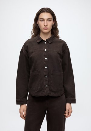 Femme aux longs cheveux ondulés portant une veste en jean marron foncé boutonnée et un pantalon assorti, debout devant un fond clair uni.