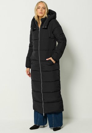 Femme portant un long manteau matelassé noir avec capuche, fermeture éclair à l'avant, mains dans les poches, associé à un jean bleu à jambes larges et des chaussures noires.