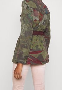 Kurtka w oliwkowym kolorze z floralnym wzorem, z paskiem w talii, w odcieniach czerwieni i z motywem liści, z wyraźnym kołnierzem.
