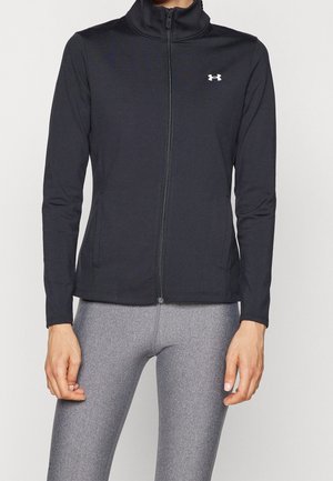 Person trägt eine schwarze Jacke mit Reißverschluss und einem weißen Logo auf der Brust sowie eng anliegende graue Leggings, steht vor einem einfarbigen Hintergrund.