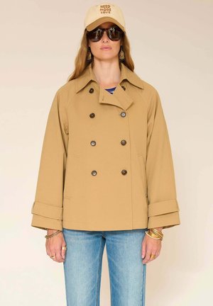 Femme portant une veste beige croisée, un jean bleu clair, une casquette beige avec le texte "NEED MORE LOVE", des lunettes de soleil foncées et des bijoux en or.