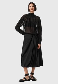 Blusa nera trasparente a maniche lunghe con scollatura arricciata, abbinata a una gonna nera con dettagli drappeggiati e sandali pianeggianti con cinturini alla caviglia.